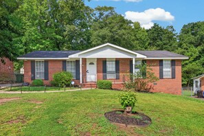 A two-story brick house with a front porch, a small front yard, and a tree in the center.