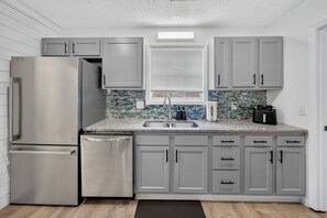 A kitchen with grey cabinets, a stainless steel refrigerator, a dishwasher, a sink, and a window with blinds.