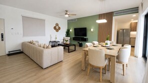 A modern living room with a sofa, dining table, chairs, a flat-screen TV, and a kitchen area visible through an open door. - Coral Juliette •Luxury PH in Cocotal • Pool • BBQ (Punta Cana)