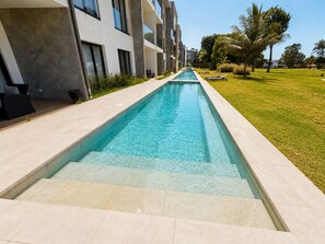 En swimmingpool med ett klart, blått vatten, omgiven av en gångbana med kakelplattor, och intilliggande en modern byggnad med balkonger.