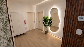 A hallway with a wooden floor, a potted plant, a mirror with a wooden frame, and a door with a fire extinguisher. - Coral Juliette •Luxury PH in Cocotal • Pool • BBQ (Punta Cana)