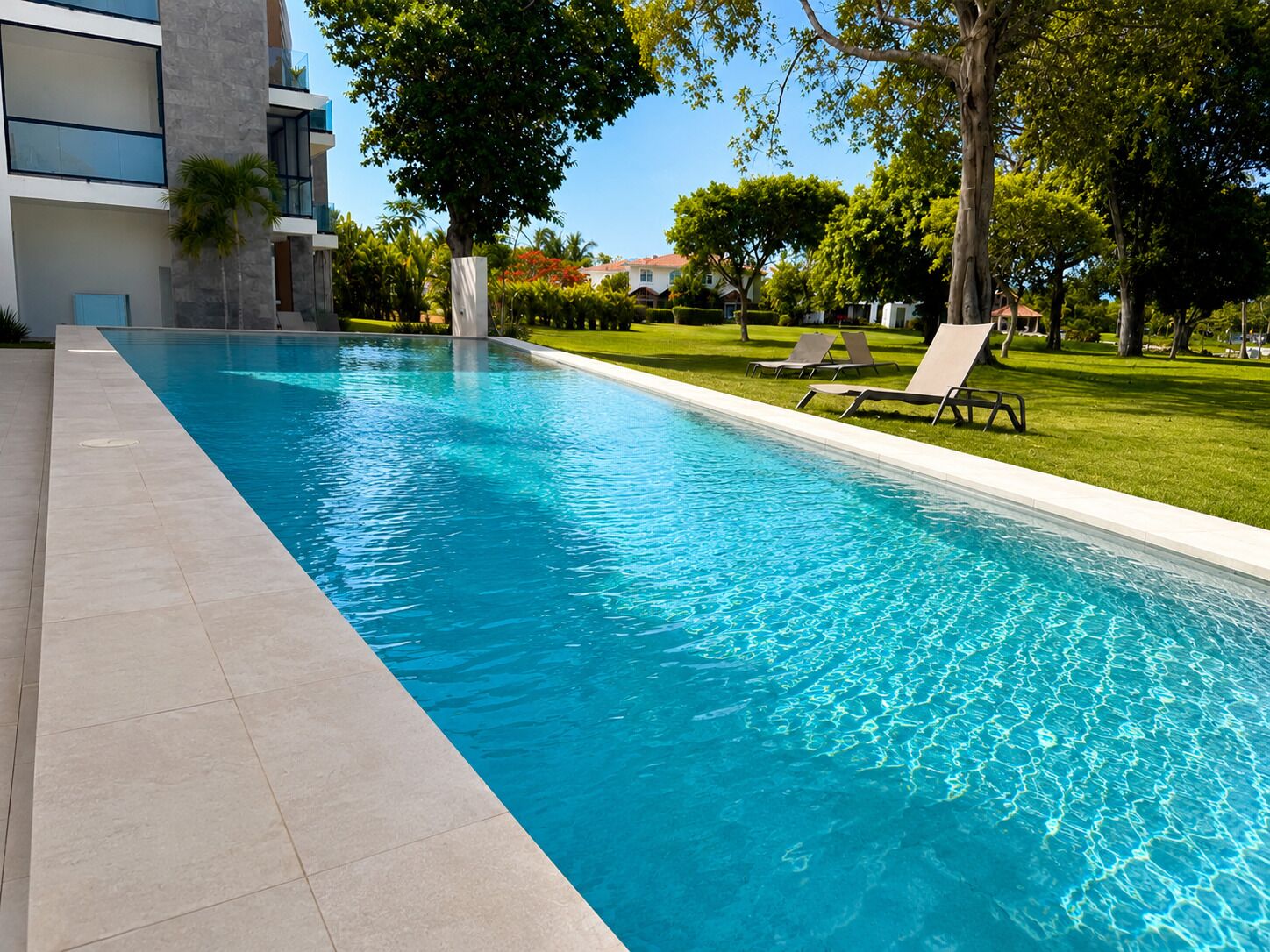 Piscina con agua azul cristalina, rodeada por un solárium con baldosas y una zona ajardinada con sillas de descanso.