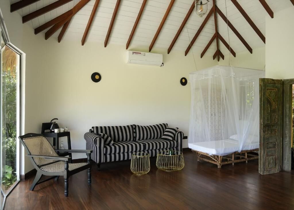 Une pièce avec un plafond en bois, un canapé à rayures noires et blanches, un fauteuil en osier, un lit doté d’une moustiquaire et une petite table avec une lampe.