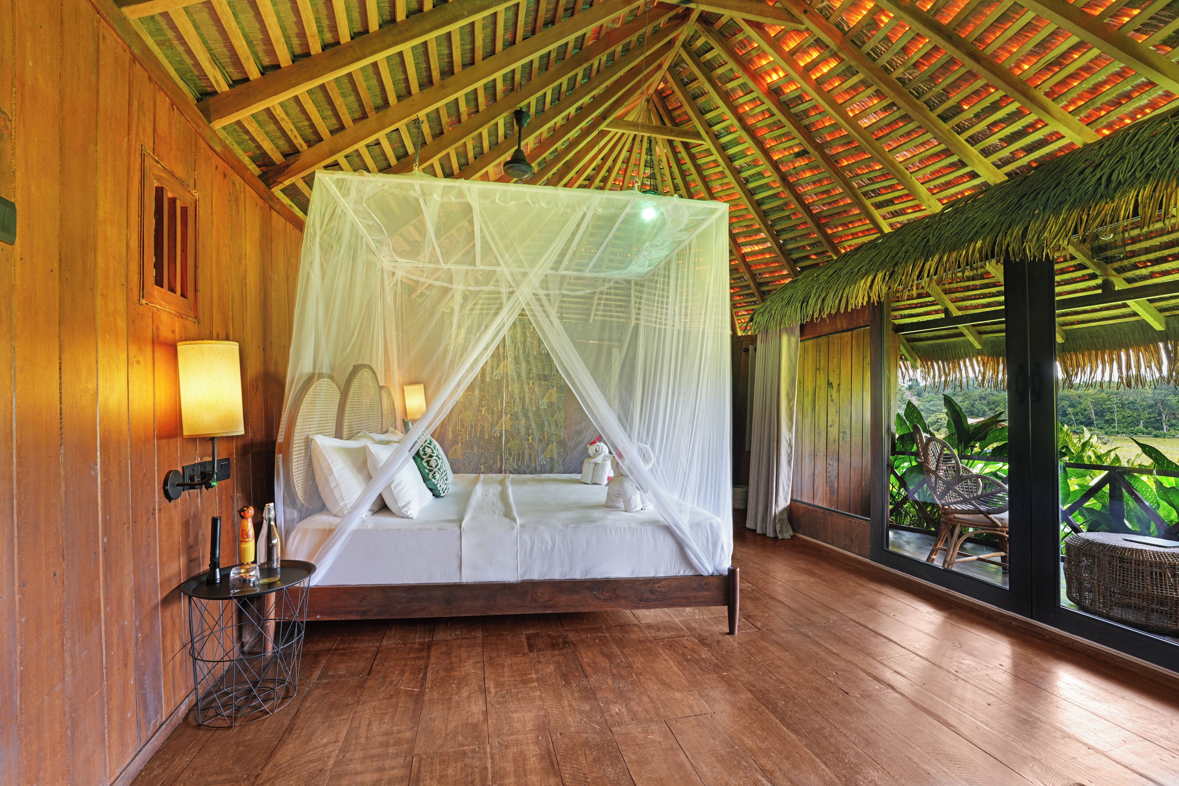 A room with a wooden floor, a bed with a canopy, a nightstand, a lamp, and a view of greenery outside.