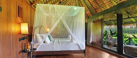 Una habitación con piso de madera, una cama con dosel, una mesita de noche, una lámpara y vistas al verdor exterior.