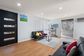 Una sala de estar moderna con un sofá negro, un televisor de pantalla plana y un amplio ventanal con persianas.