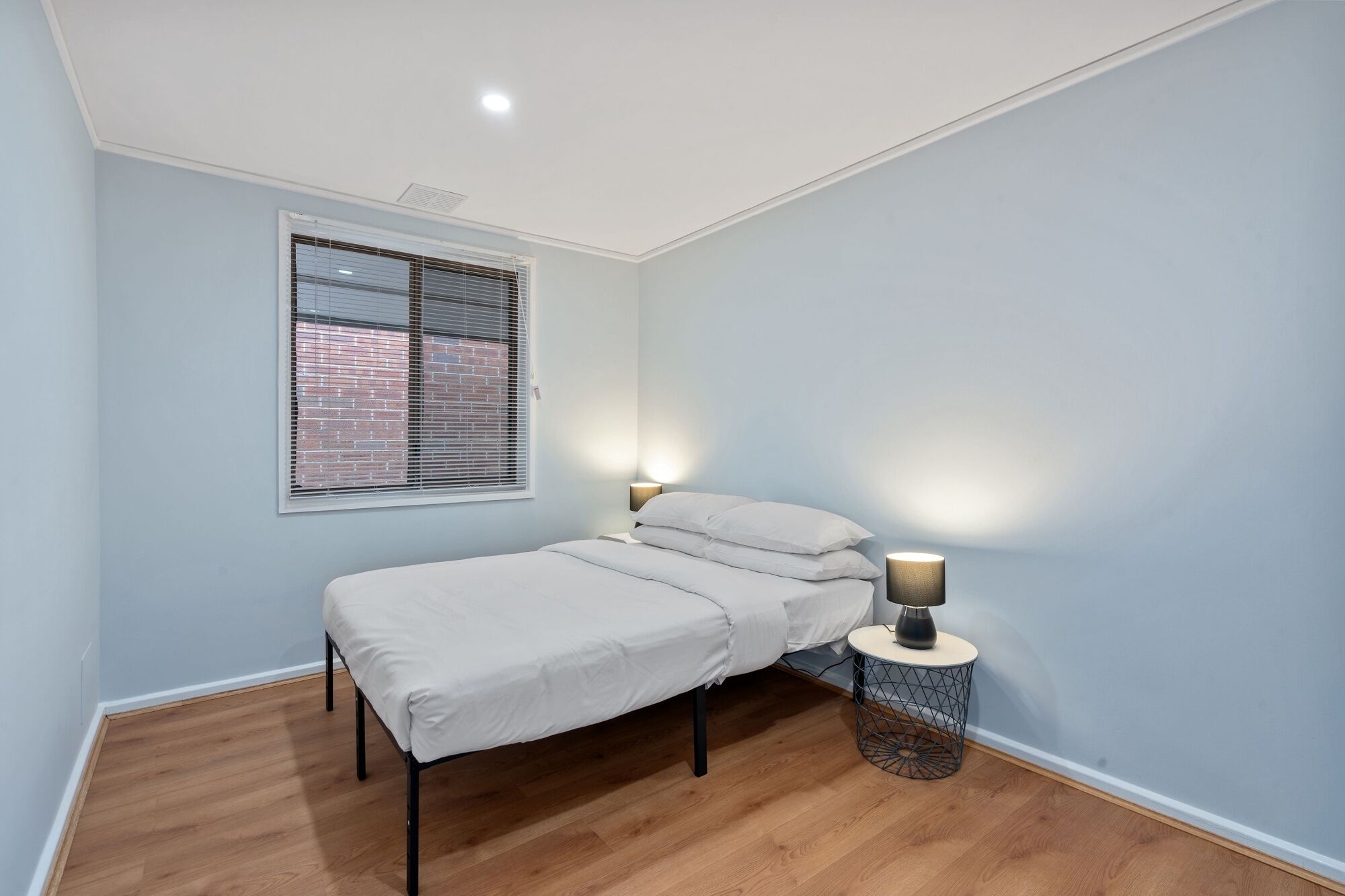 Ein Schlafzimmer mit einem Einzelbett, einem Fenster mit Jalousien, einer Nachttischlampe und einem runden Beistelltisch.
