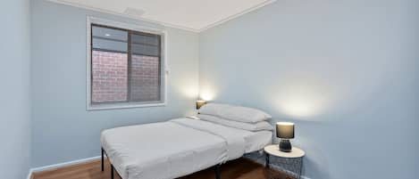 Ein Schlafzimmer mit einem Einzelbett, einem Fenster mit Jalousien, einer Nachttischlampe und einem runden Beistelltisch.