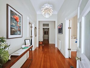 A hallway with wooden flooring, a white ceiling, and a chandelier. There are framed pictures on the walls and a plant on a table.