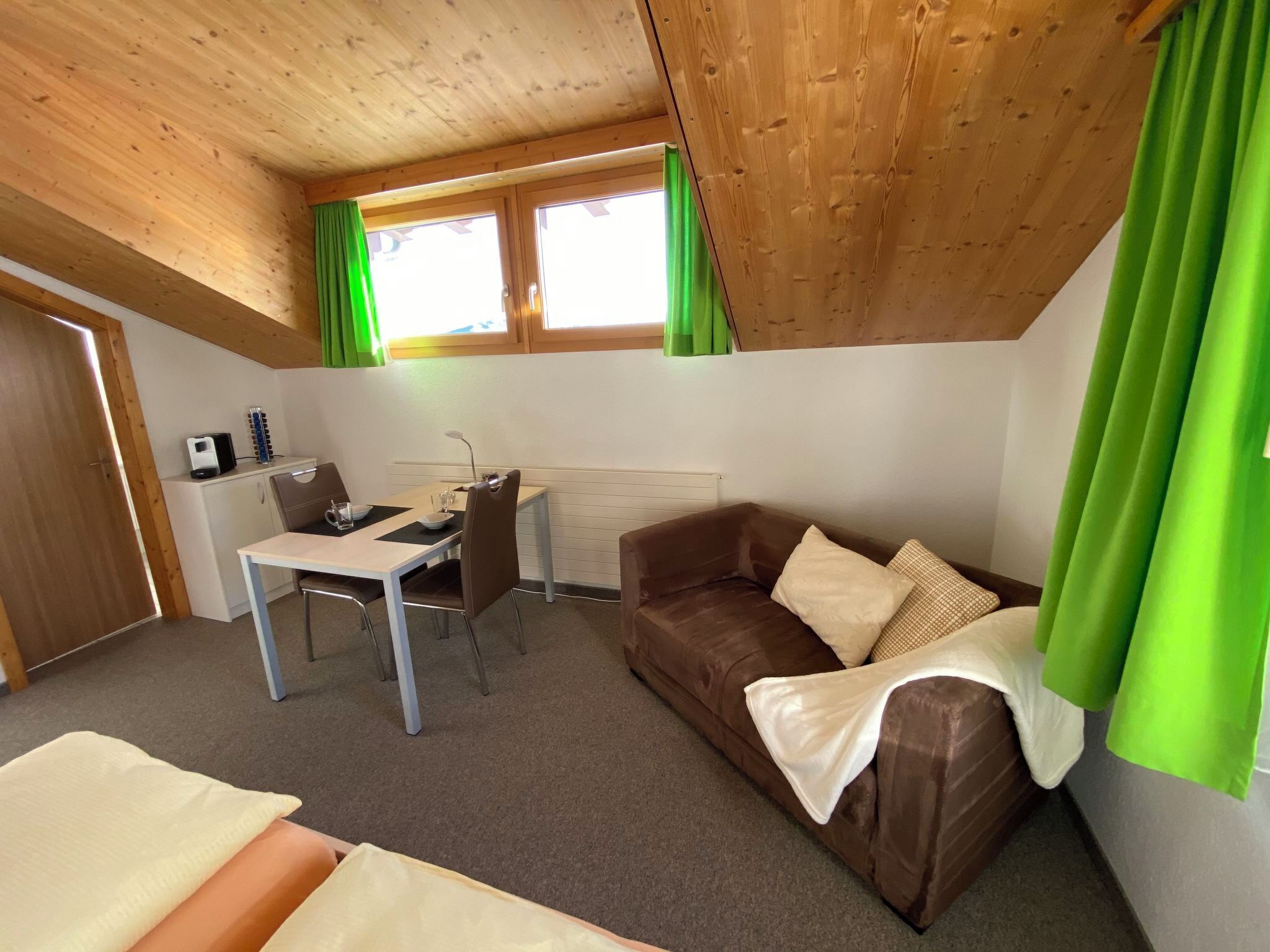 Et lille, hyggeligt rum med træloft, en brun sofa med puder, et lille skrivebord med stol og et vindue med grønne gardiner.