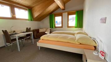 A small, cozy room with a bed, a desk, and a chair. There are two windows with curtains, and the room has a sloped ceiling.