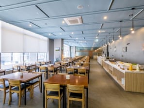 Un comedor amplio con varias mesas y sillas, una barra larga con alimentos y ventanas grandes que permiten la entrada de luz natural.