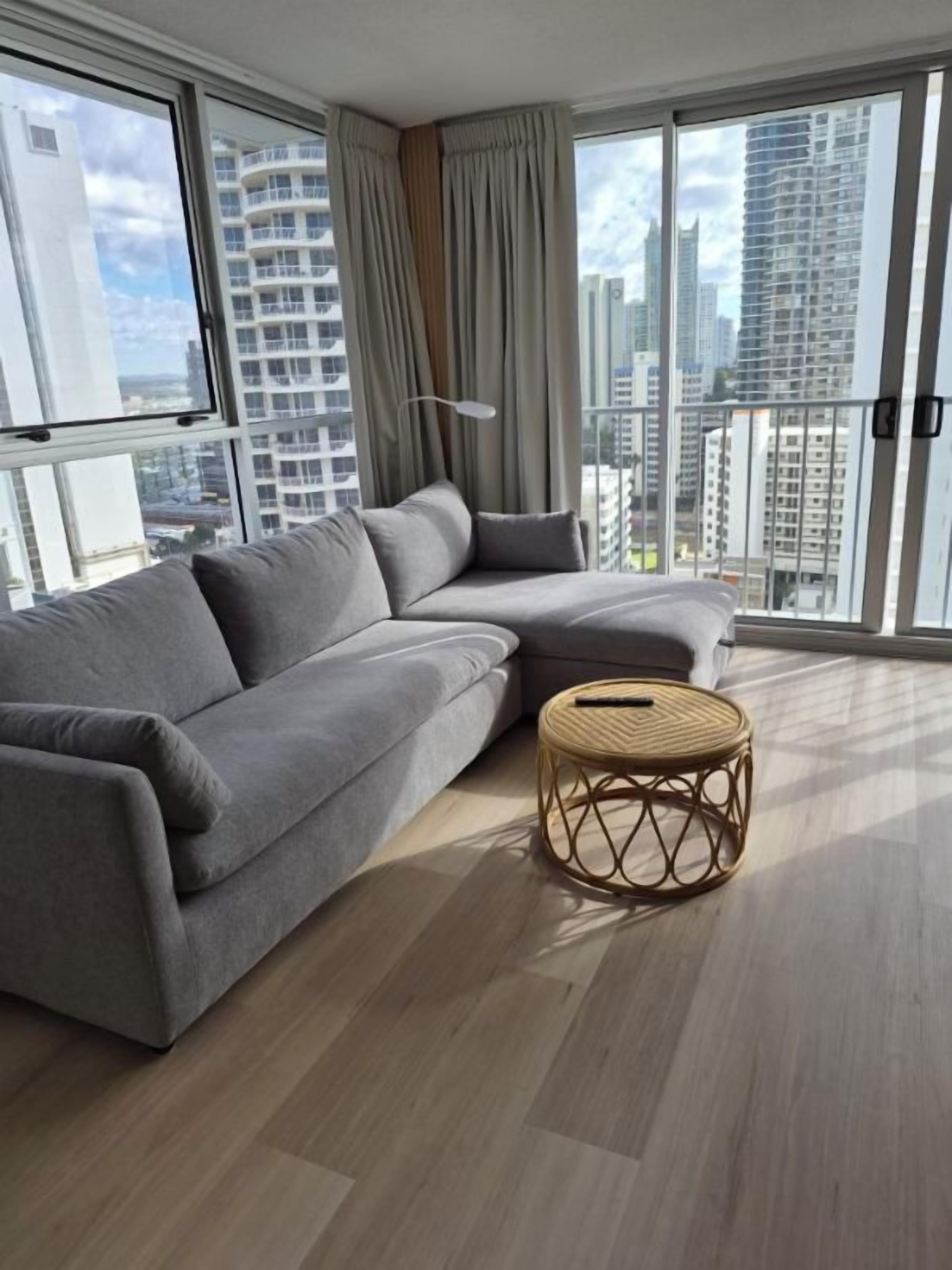 Una sala moderna con un sofá seccional gris, una mesita auxiliar de mimbre redonda y amplios ventanales de piso a techo con vistas a la ciudad.