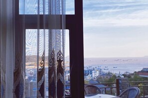 A view through a window with ornate curtains, a balcony with a chair, and a distant cityscape with buildings and boats. - Shengsi hairongju seascape homestay (Shengsi)