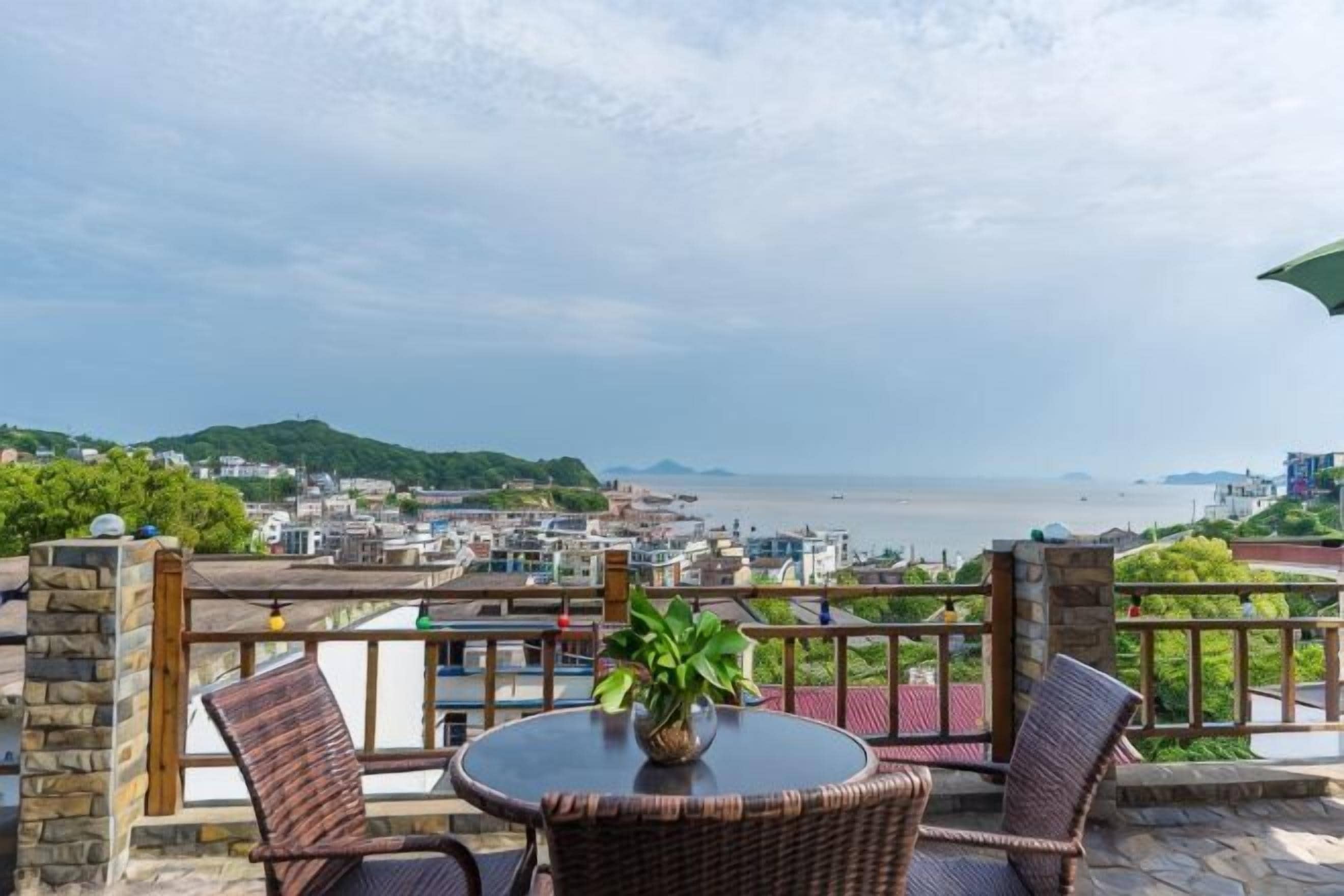 A terrace with wicker furniture and a potted plant, overlooking a coastal cityscape.