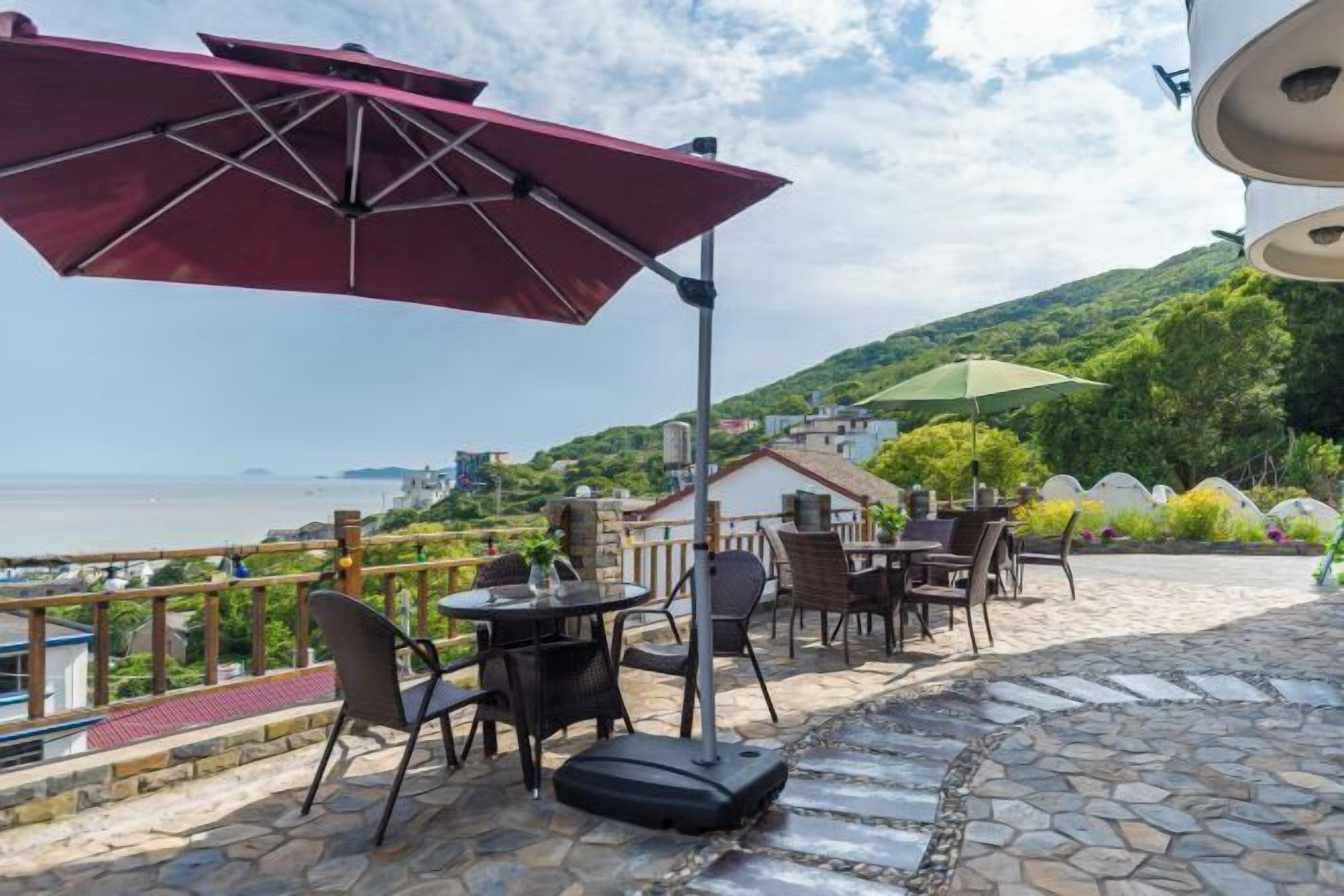 An outdoor patio with tables and chairs, a red umbrella, and a view of the sea.