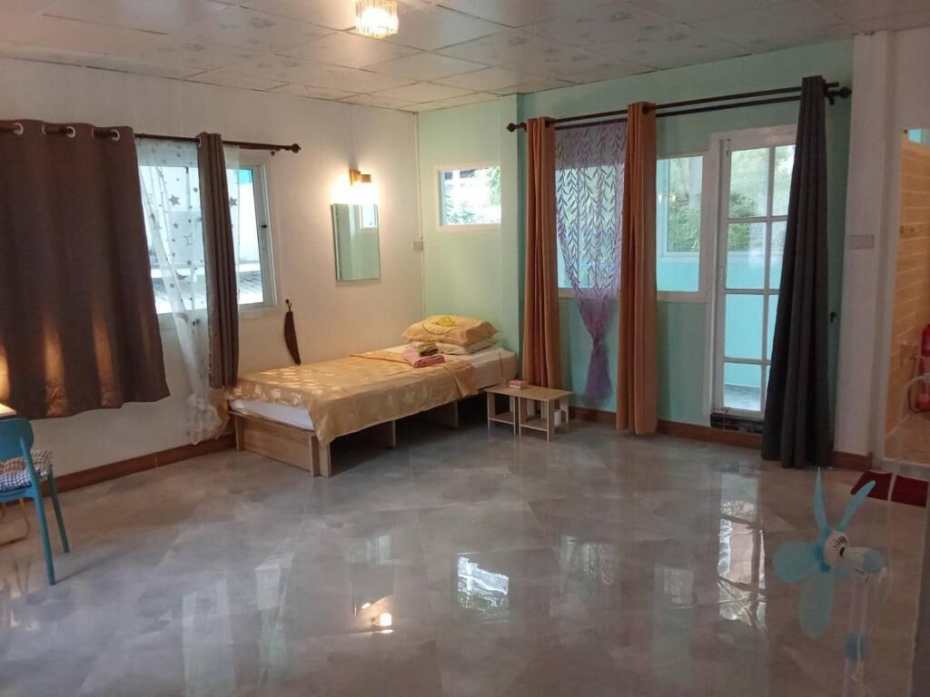 Una habitación con cama, una mesita y una silla. Se observan ventanas con cortinas, una puerta y una lámpara en el techo.