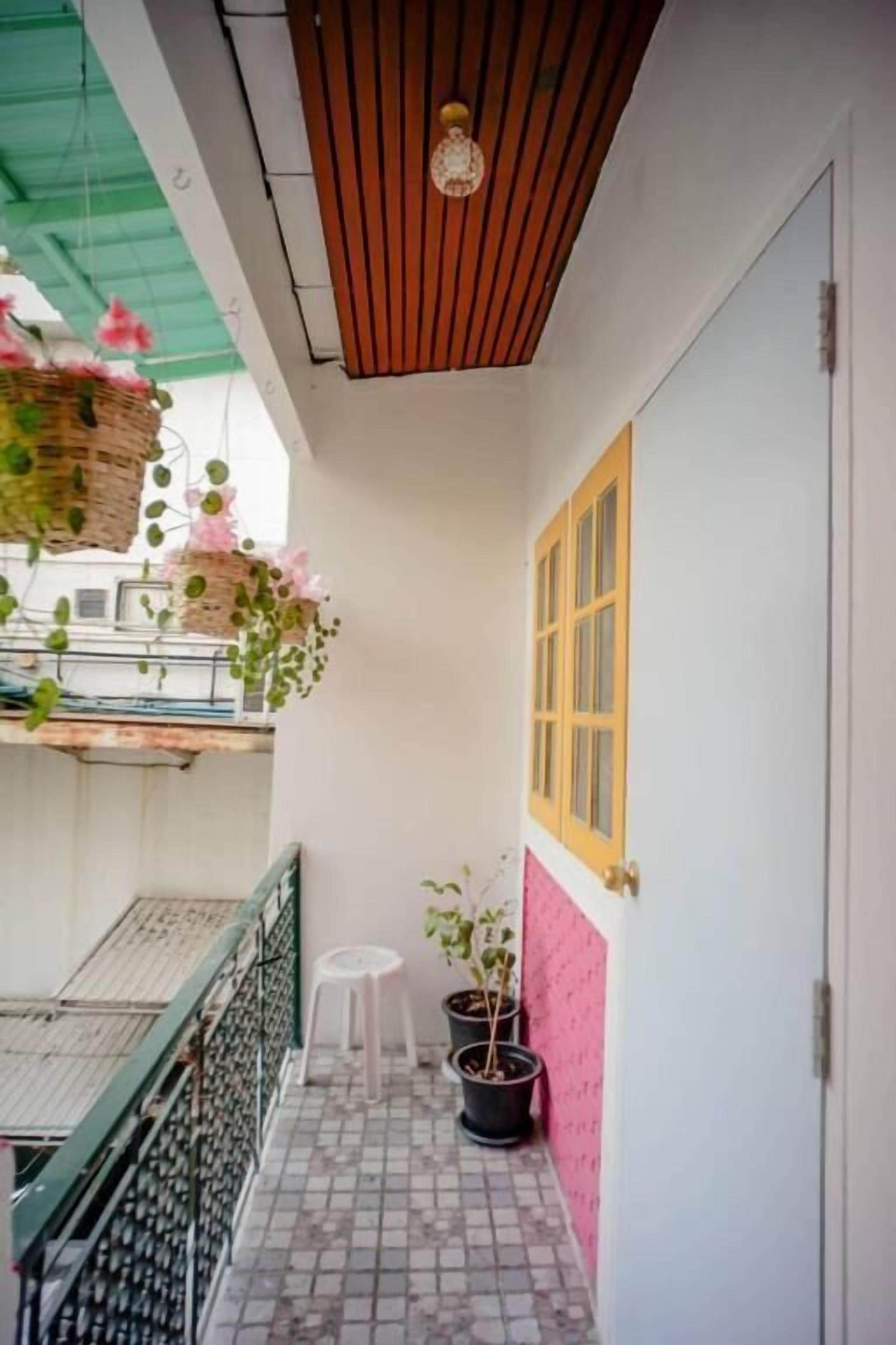 A narrow balcony with a tiled floor, a white door with a yellow window, and hanging plants.