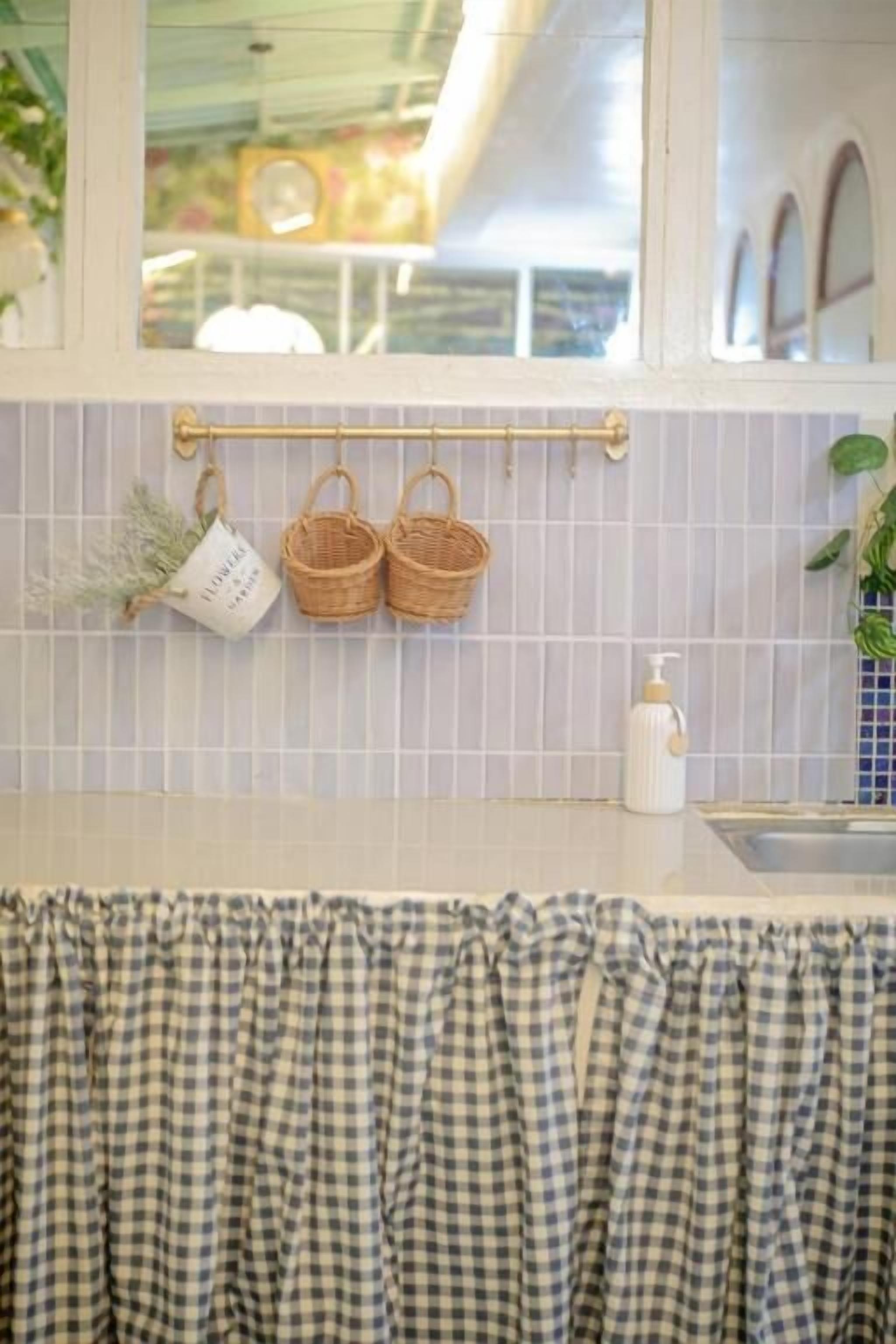 A kitchen with a checkered curtain, a hanging rack with baskets, and a window with a view of the outdoors.