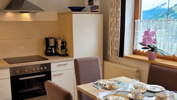 A compact kitchen with a stove, oven, and a coffee maker. There is a dining table set with plates, glasses, and cutlery. A window with a view of mountains is visible. - Zuppinger by Interhome (Mayrhofen)