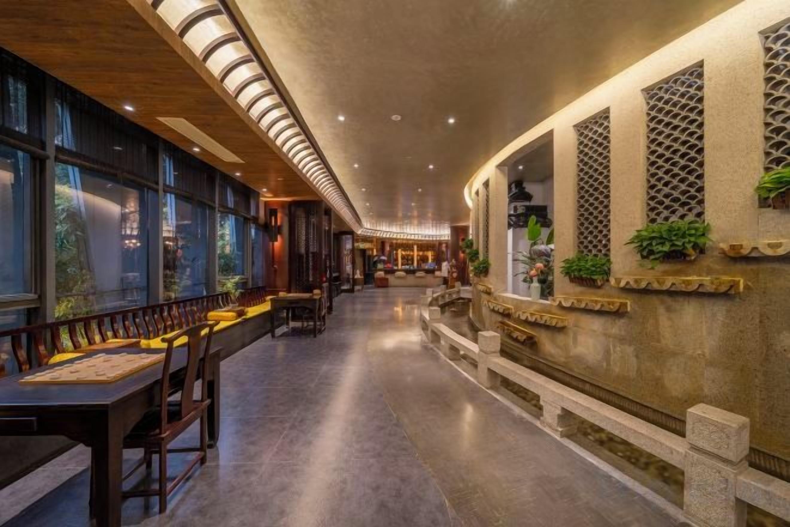 A spacious corridor with a long table and chairs, a seating area with cushions, and a wall with decorative elements and plants.