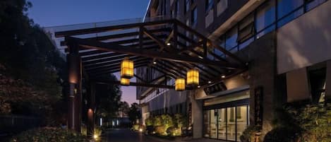 Una entrada de hotel con un paseo techado, iluminada por faroles colgantes, rodeada de follaje.