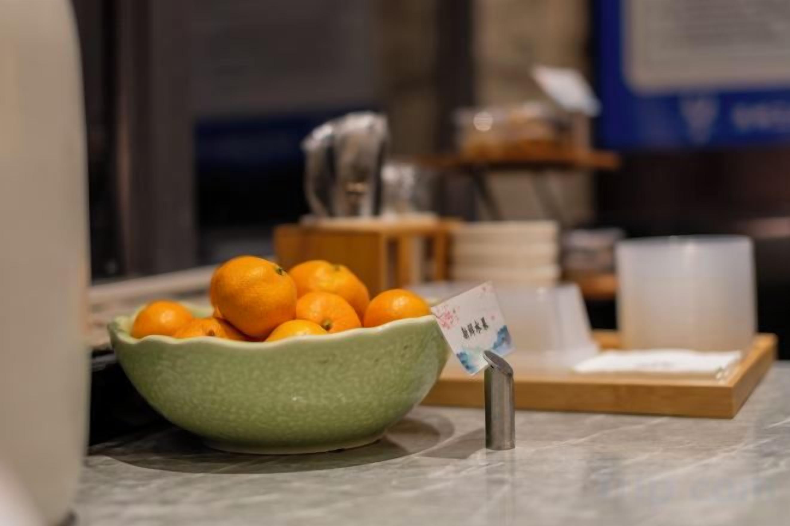 A green bowl filled with oranges on a marble countertop, with a small sign next to it.