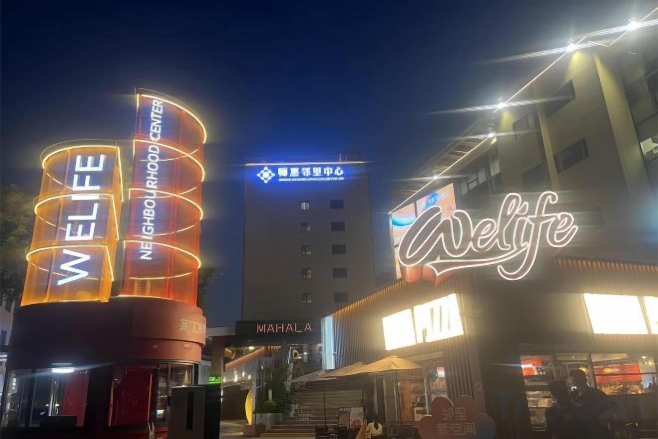 A well-lit urban street scene with illuminated signage for a hotel named 'WELIFE' and a building with a sign in a foreign language.