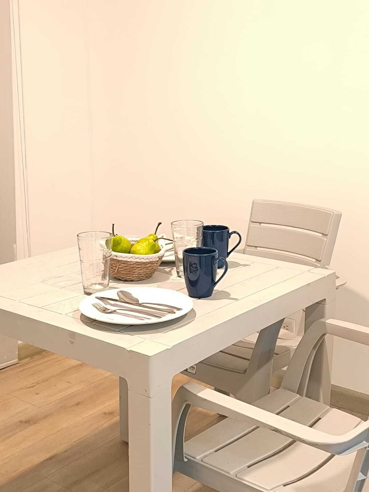 A white dining table with a plate, cutlery, a glass, a blue mug, and a basket of lemons.