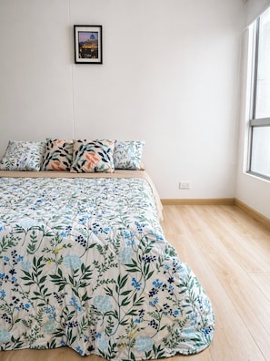 Um quarto com colcha estampada de flores, um quadro na parede e uma janela que proporciona luz natural.