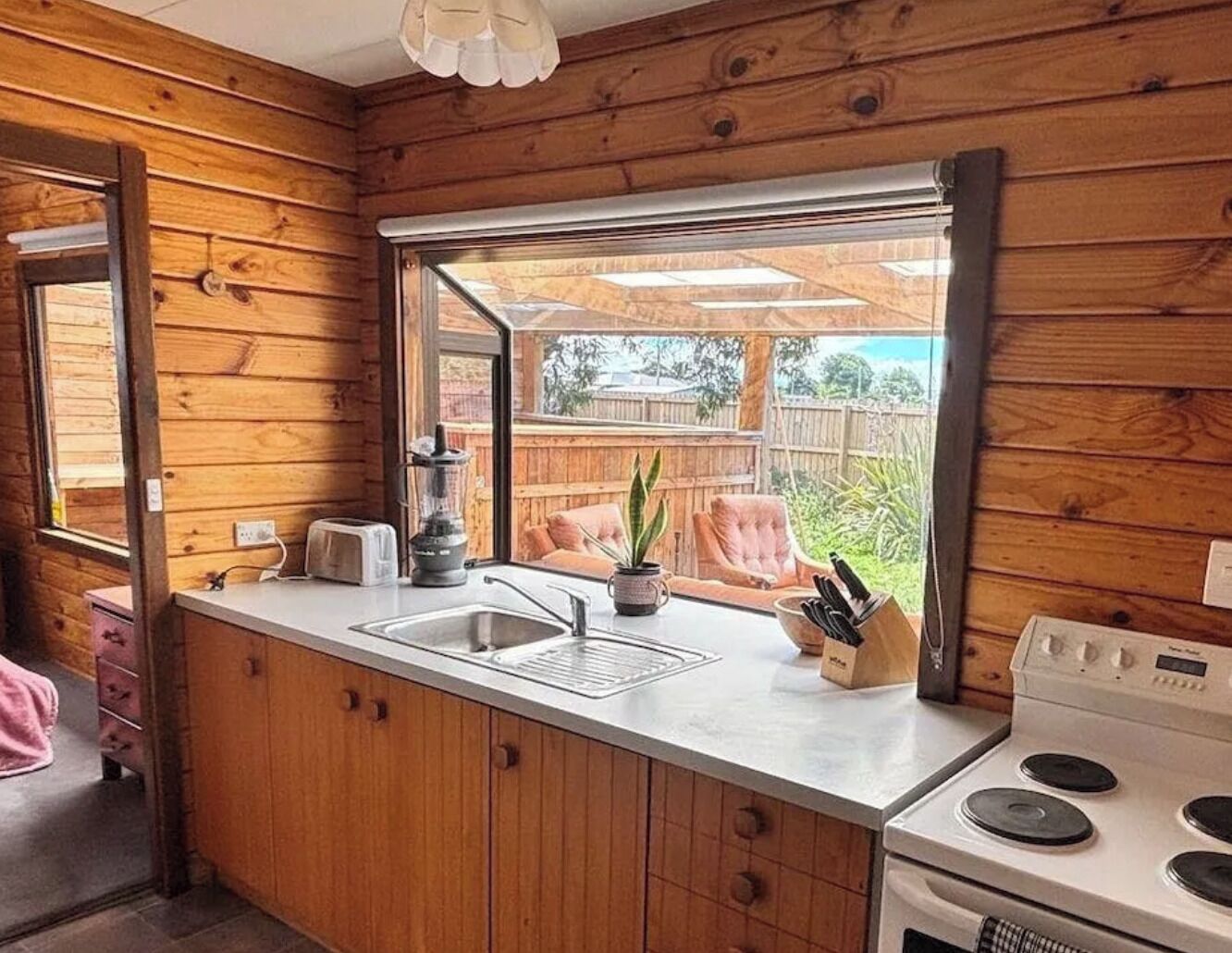 A compact kitchen with wooden walls, a window, a sink, a toaster, a stove, and a small countertop area.