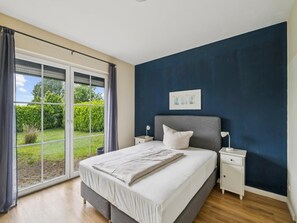 A bedroom with a large bed, a nightstand, a bedside table, and a view through a sliding glass door to a garden. - Cozy holiday apartment with terrace and garden. (Bastorf)