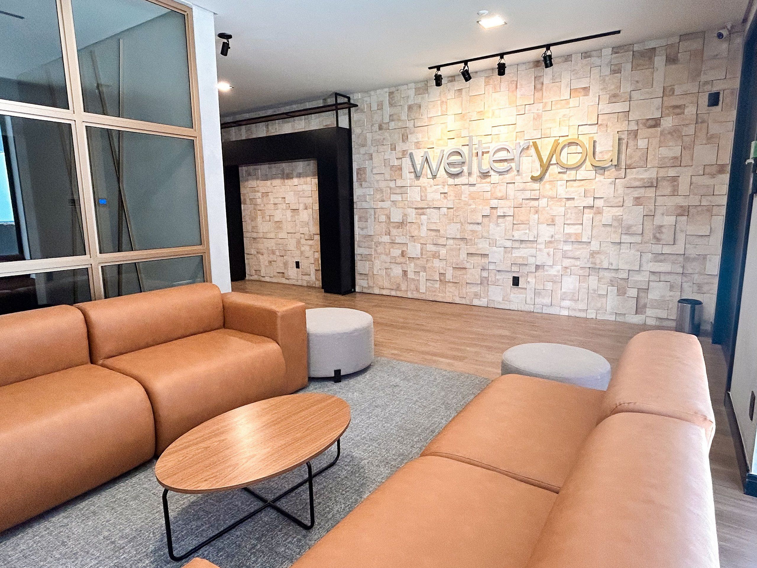 Un interior moderno con un sofá modular, una mesa redonda de madera y una pared con diseño de piedra que incluye la señalización 'welteryou'.