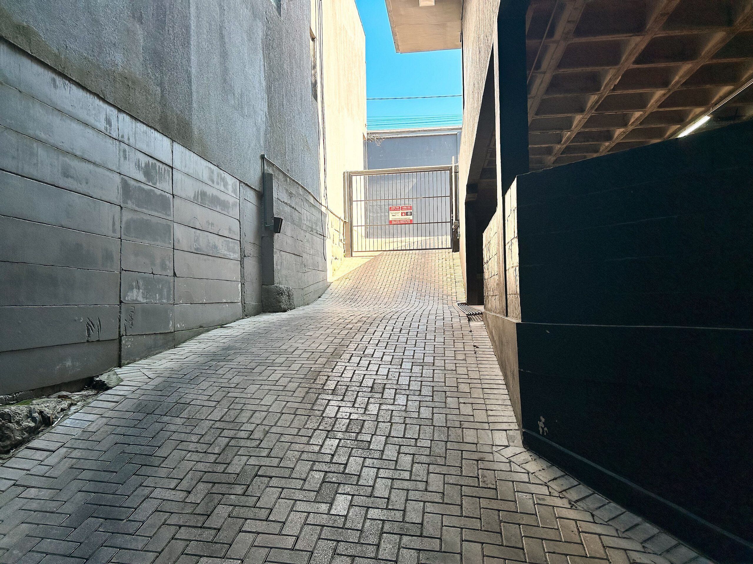 Un estrecho callejón con piso de ladrillo, una reja metálica y un muro de hormigón.