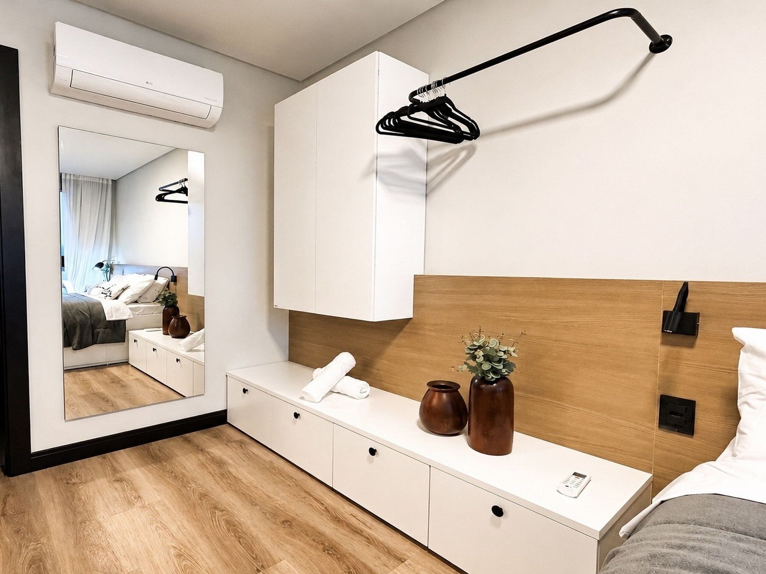 Habitación de hotel moderna con cabecera de madera, una mesita de noche con un florero y un control remoto, un espejo grande y una lámpara colgante.