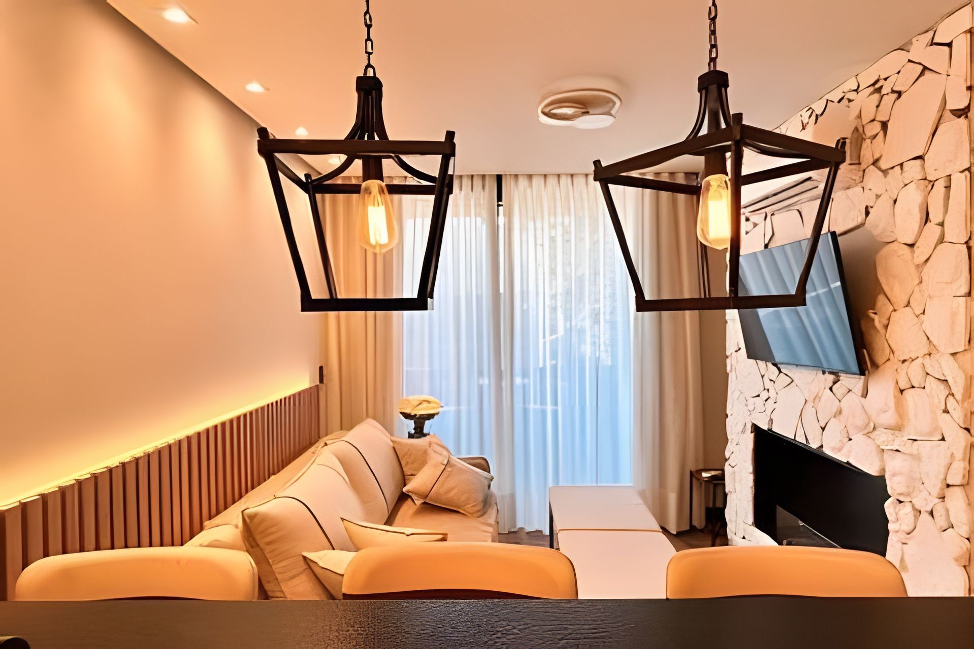 A modern living room with a sofa, a flat-screen TV mounted on a stone wall, and pendant lighting.