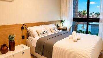 Habitación de hotel con cama, mesita de noche, lámpara y vistas amplias de la ciudad.
