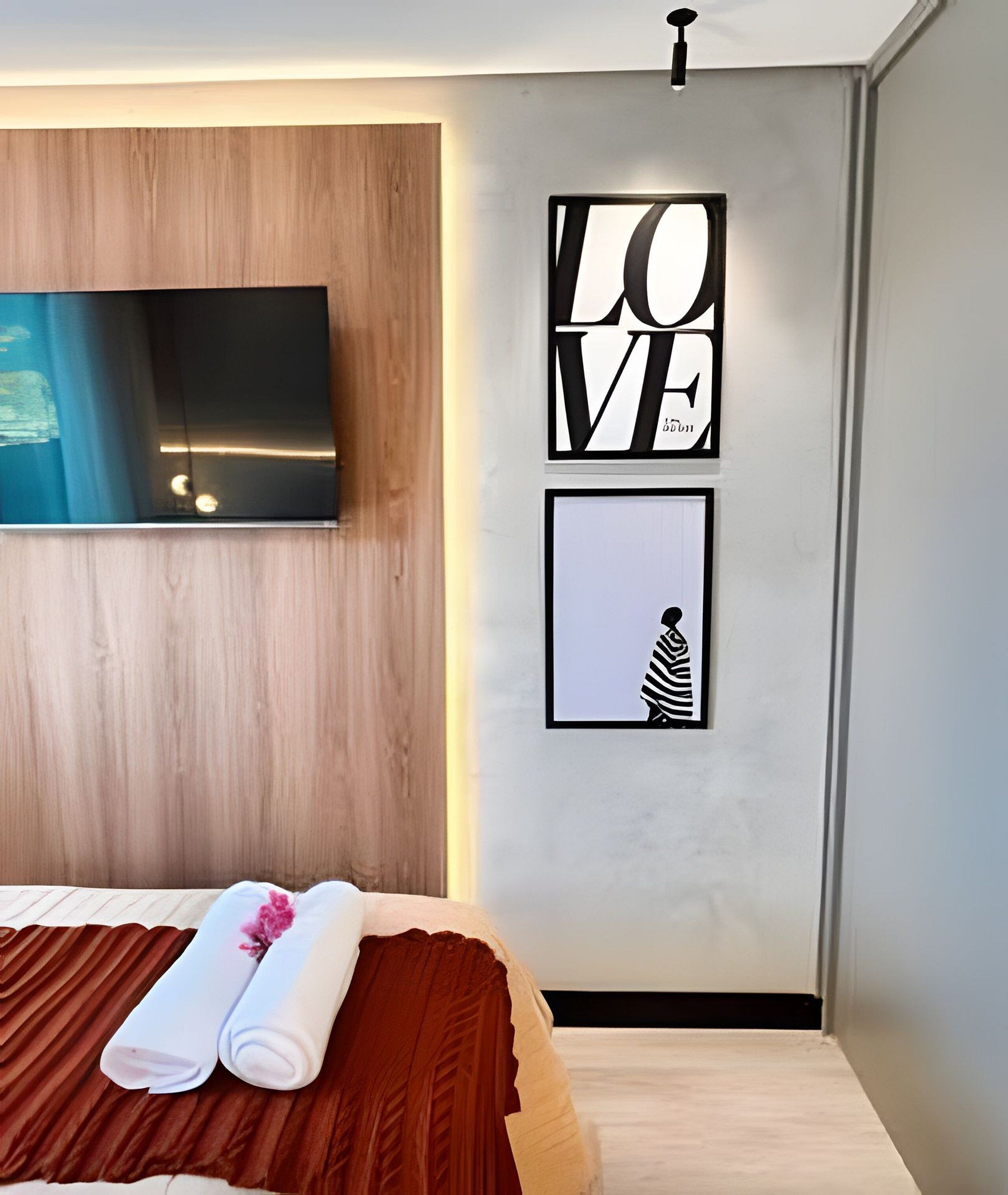A modern bedroom with a wooden headboard, a flat-screen TV mounted on the wall, and a framed artwork with the word 'LOVE'.