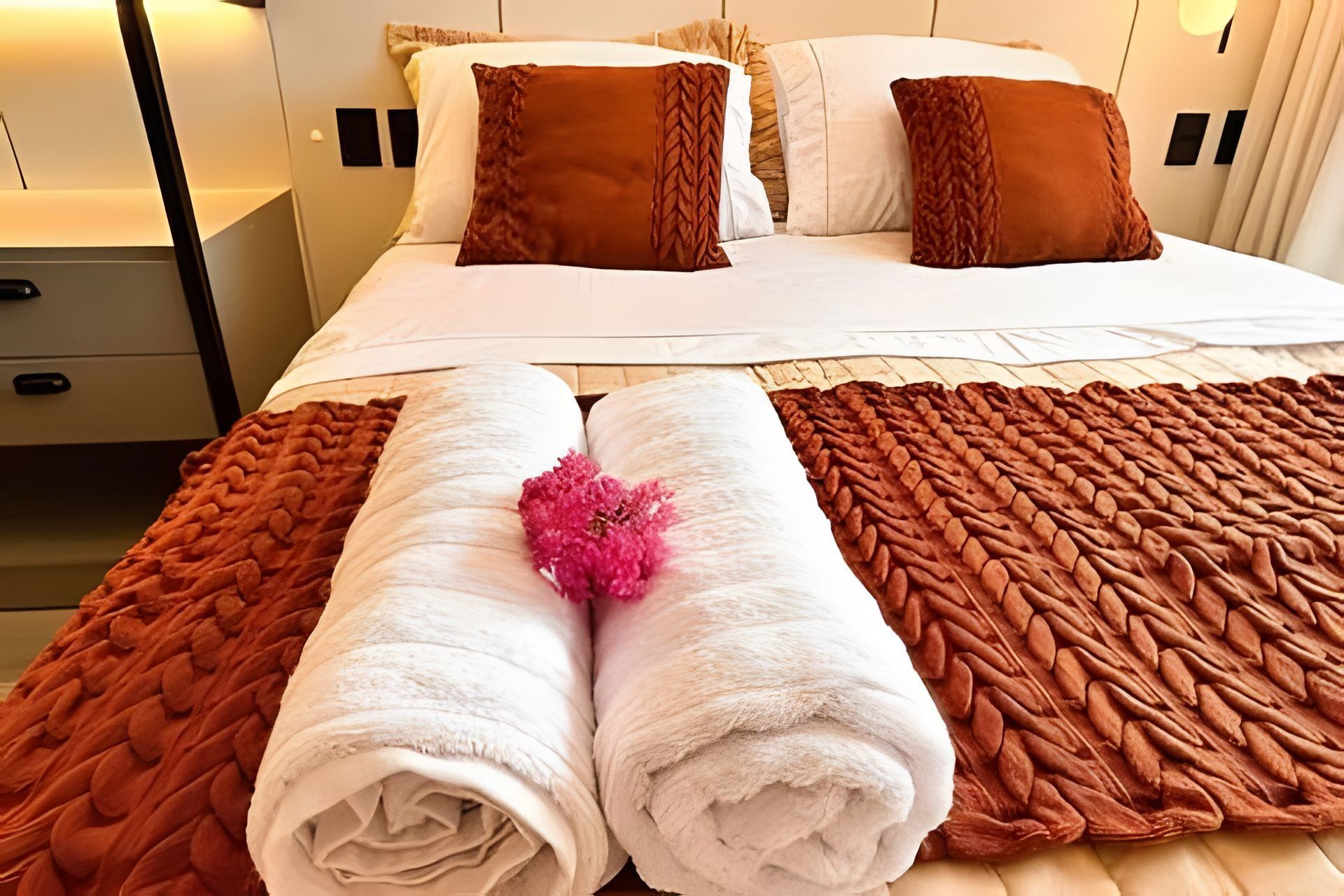 A neatly made bed with a brown blanket and two pillows, a wooden nightstand with a drawer, and a wall-mounted lamp.