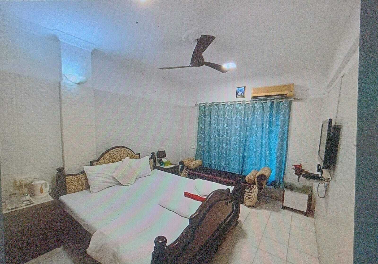 Une chambre à coucher comprenant un lit, une table de chevet, un bureau, une chaise, un ventilateur de plafond et une fenêtre avec des rideaux.