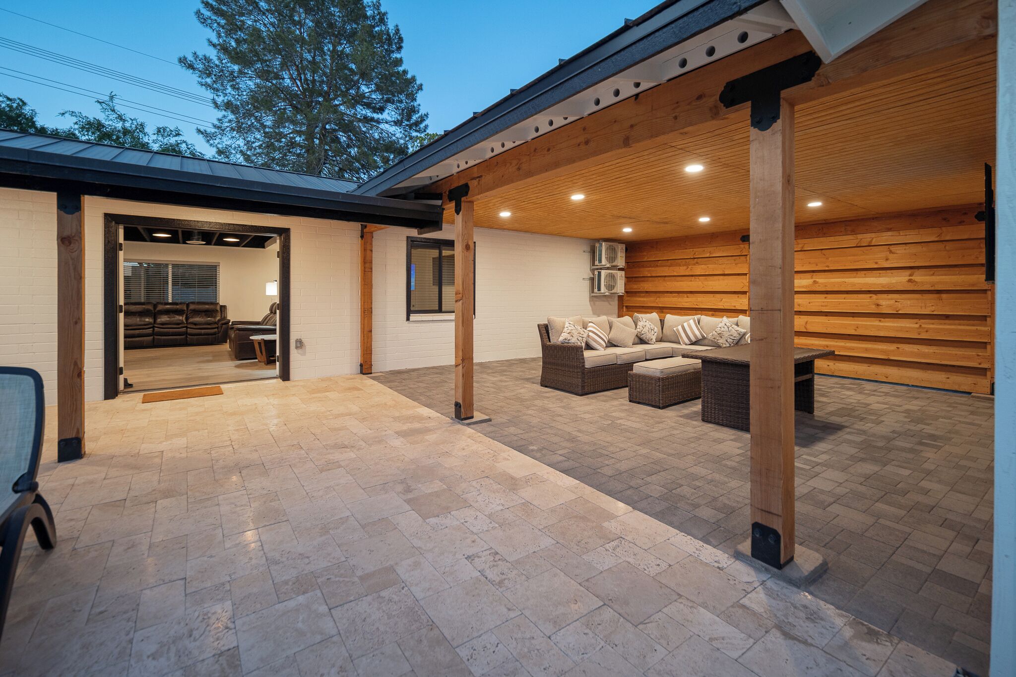 A covered outdoor patio area with a stone floor, wooden beams, and a seating arrangement.
