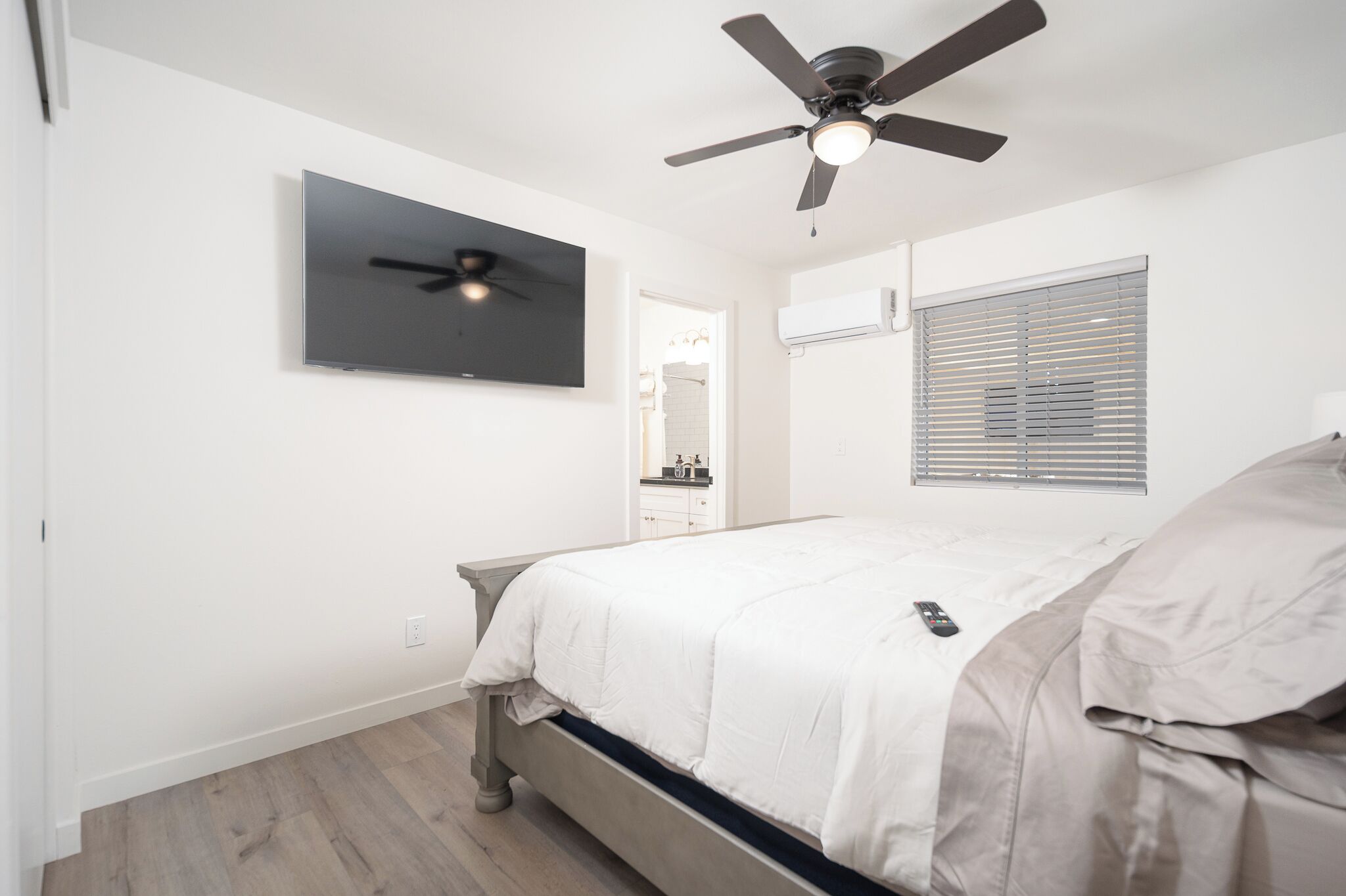 Une chambre à coucher avec un lit, un ventilateur de plafond, un téléviseur fixé au mur et une fenêtre avec des stores.