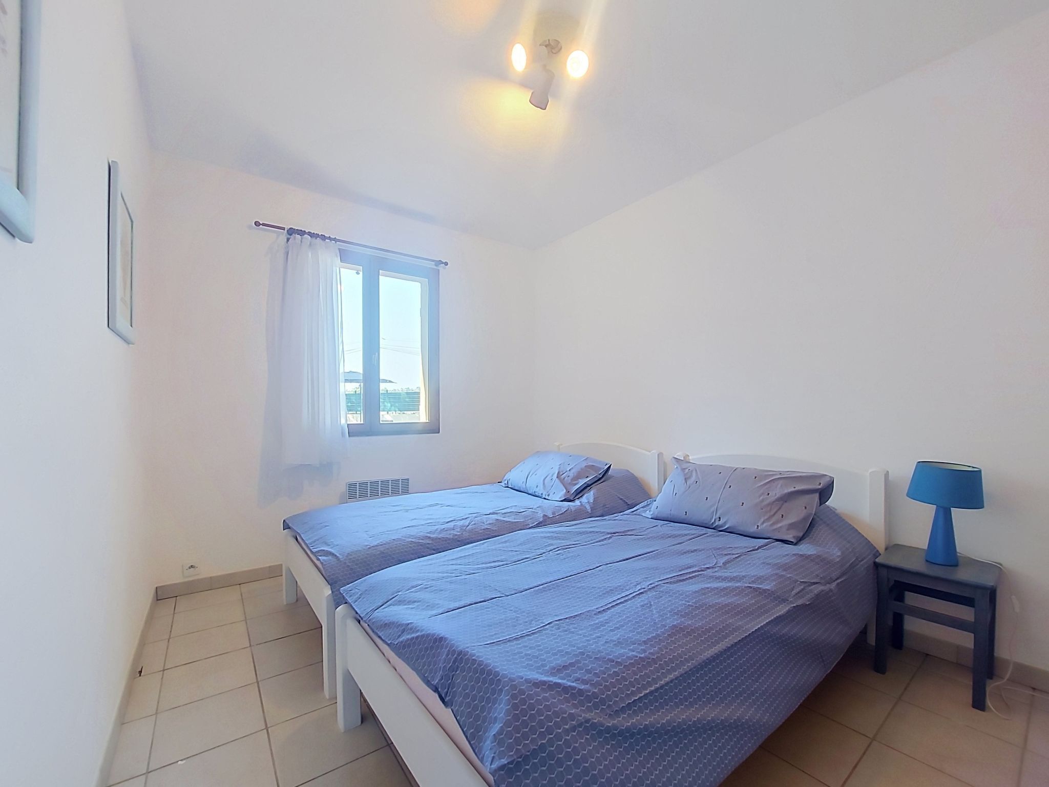 Une chambre à coucher comprenant un lit, une table de chevet, une fenêtre avec des rideaux et un luminaire fixé au mur.