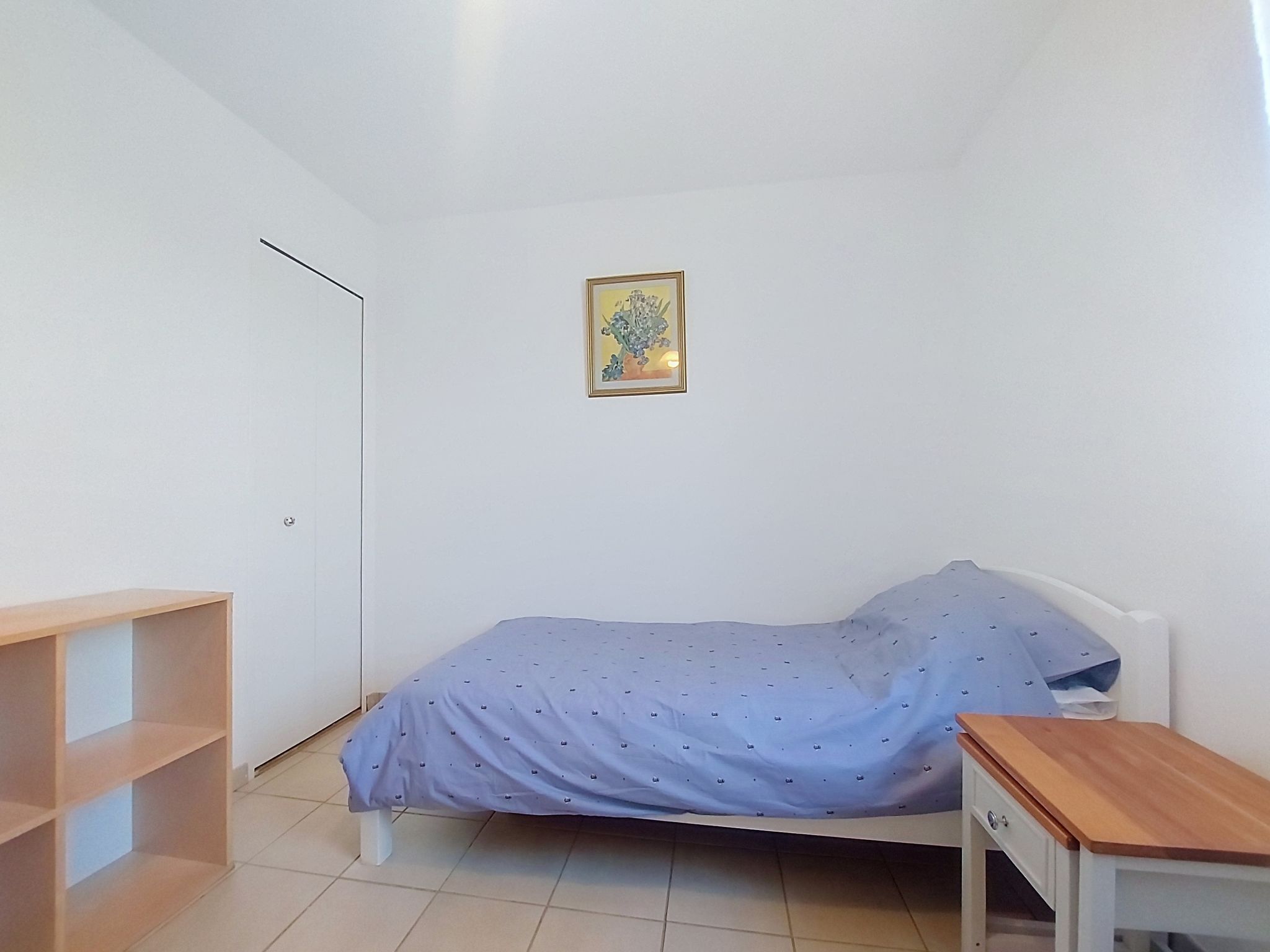 A small, sparsely furnished room with a single bed, a small table, and a wall-mounted picture.