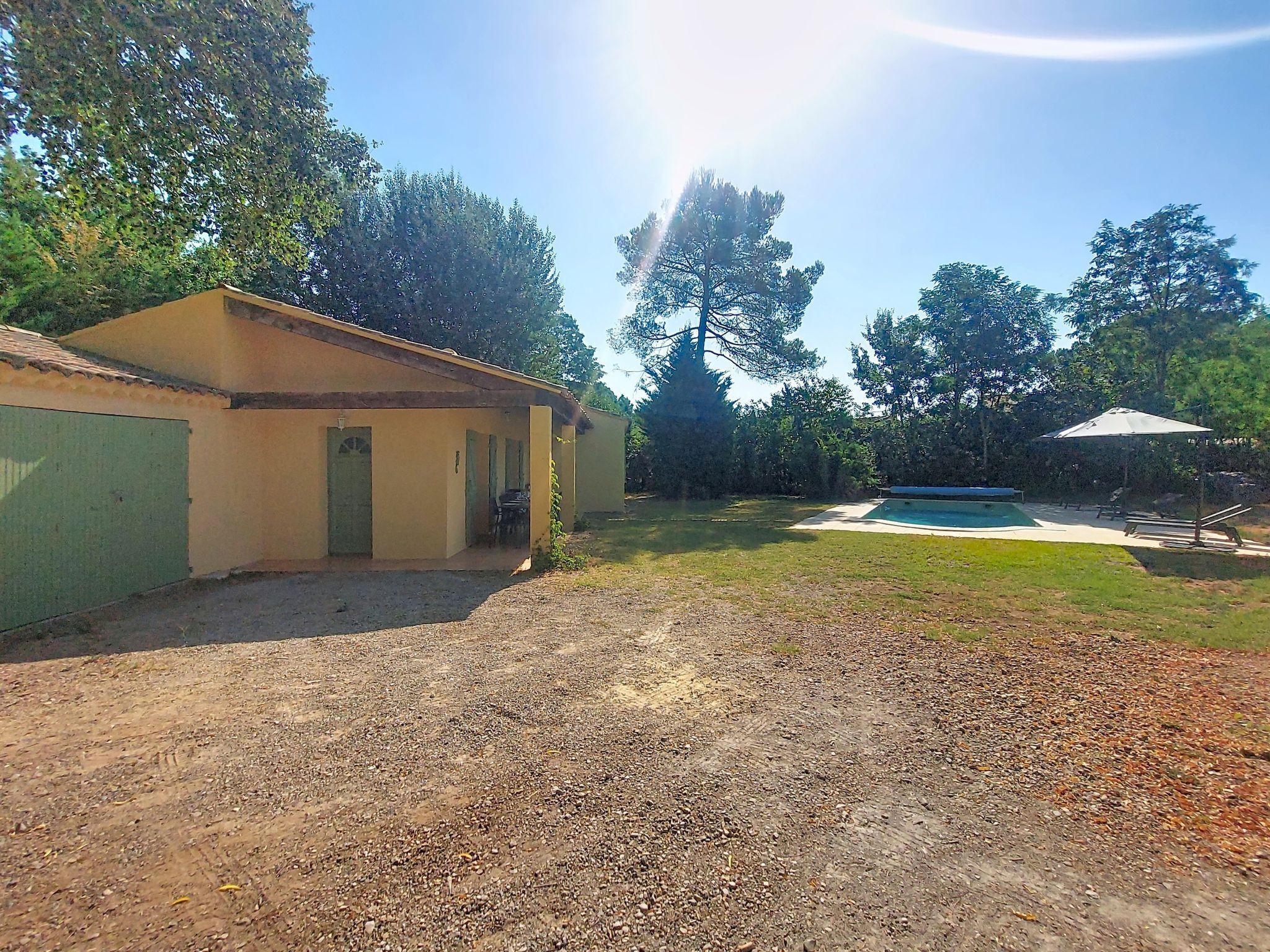 A gravel driveway leading to a building with a green door, a grassy area with a pool, and trees in the background.