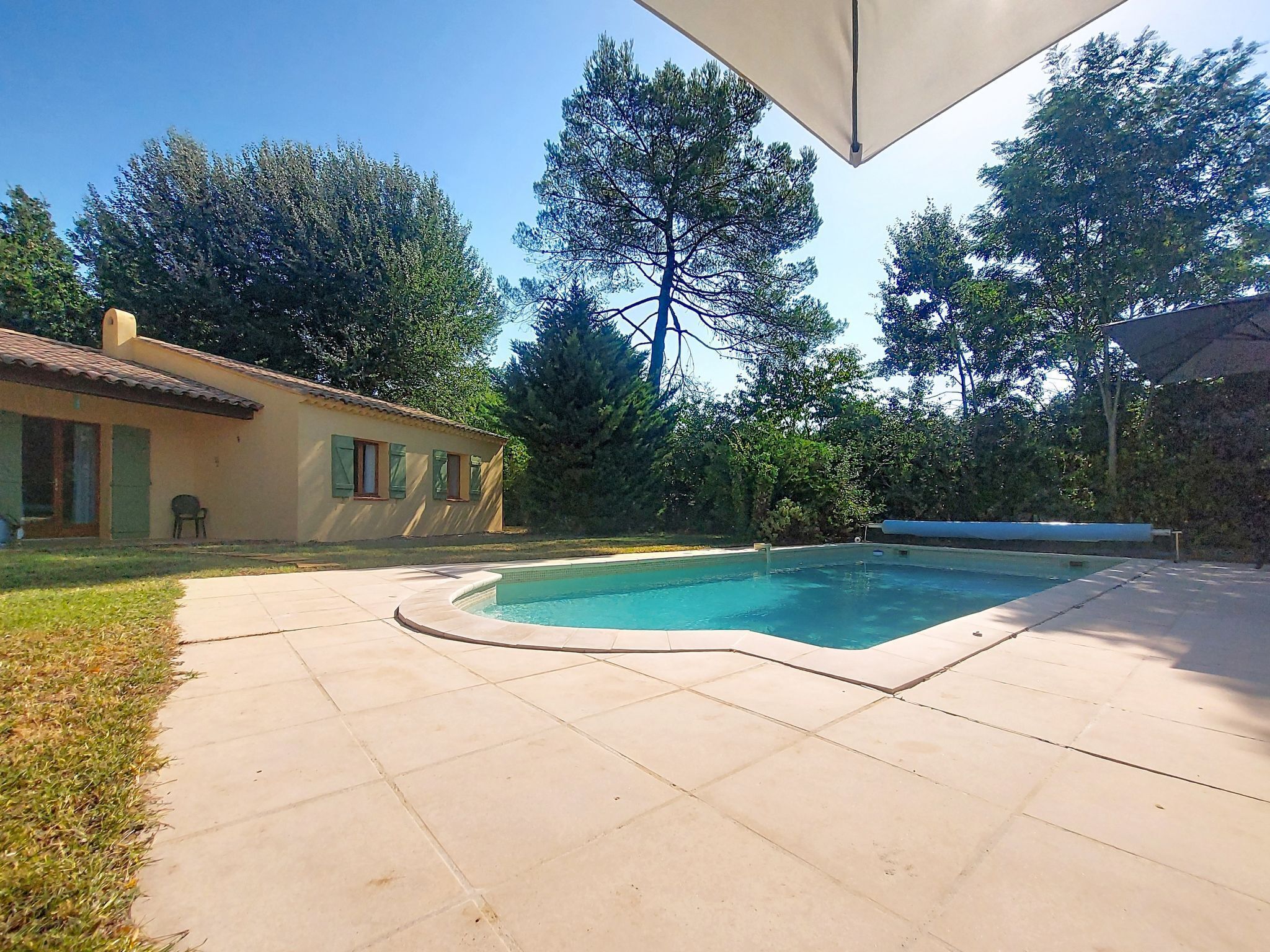 A swimming pool with a tiled deck, surrounded by trees and a building with a tiled roof.
