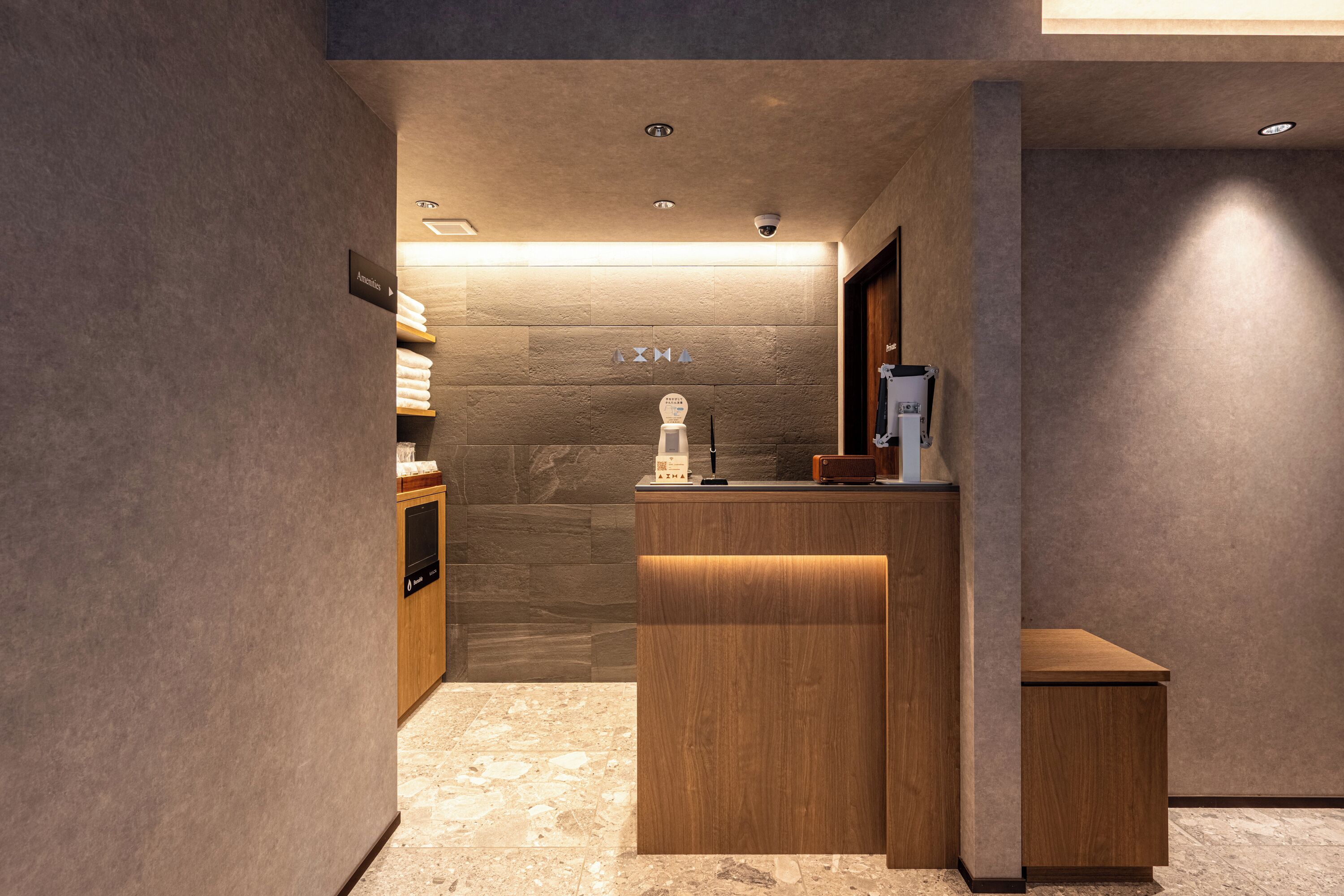 A hotel reception area with a wooden reception desk, a small seating area, and a wall-mounted sign indicating amenities.