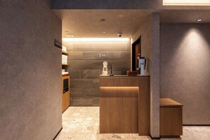 A hotel reception area with a wooden reception desk, a small seating area, and a wall-mounted sign indicating amenities. - AIMA Asakusa East (Tokyo)