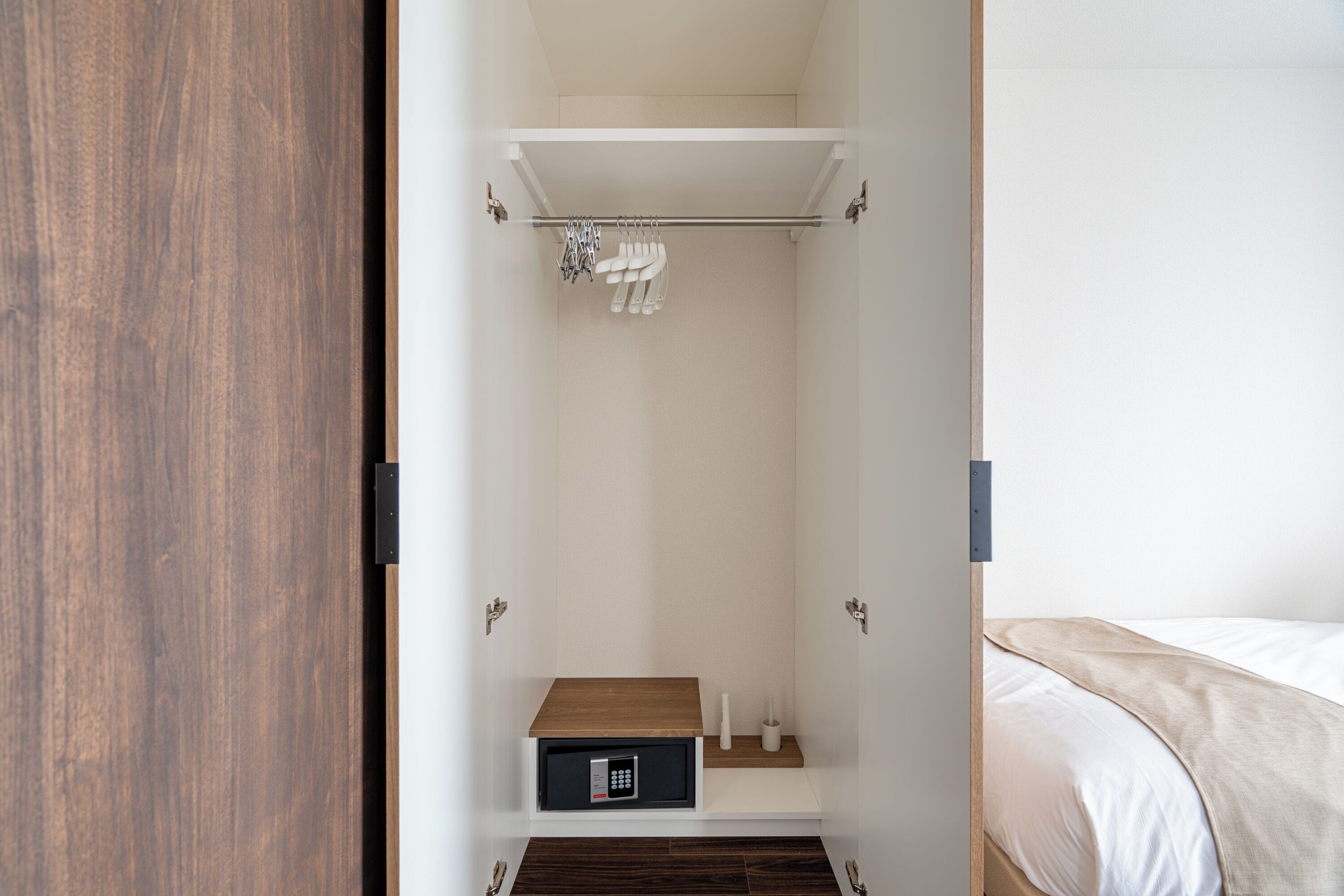 A hotel room with a wooden wardrobe, a built-in shelf with a microwave, and a bed with a beige blanket.