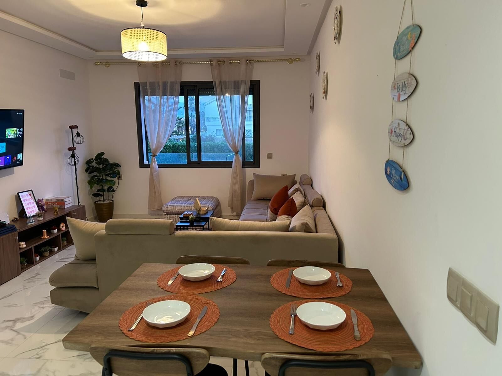 A modern living room with a sofa, dining table set for four, a TV, and a large window with curtains.